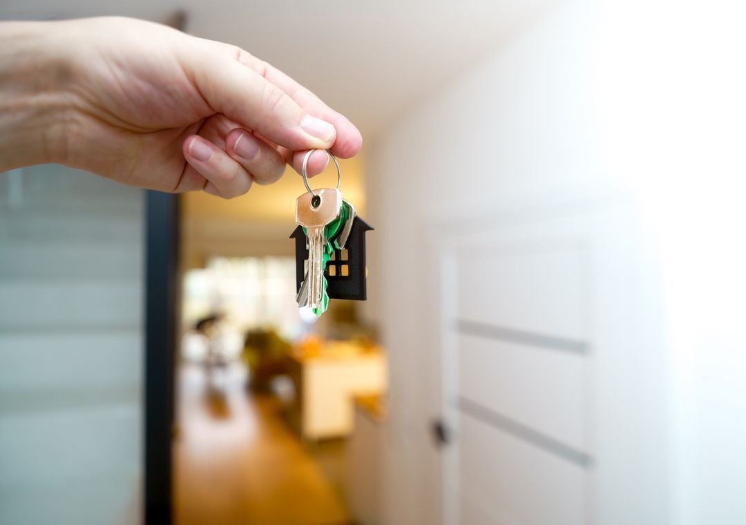 Hand holding house keys with a small house-shaped keychain near an open doorway, representing home exchange, hospitality, and slow travel.