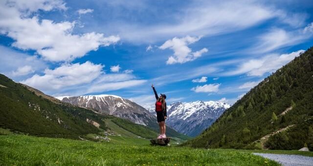 Summer landscape of Livigno in the Italian Alps, with green meadows, alpine houses and snow-covered mountains under a blue sky.