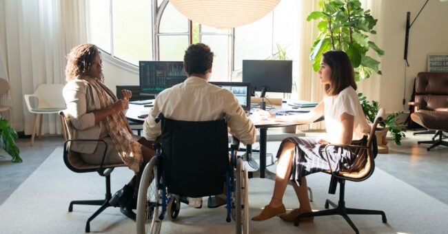 A diverse group taking part in a collaborative meeting in an accessible workplace, including a wheelchair user. Featured image for the Voluntouring.org guide about inclusive volunteering, accessibility, flexible participation and removing barriers for volunteers.