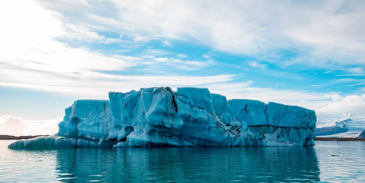 Large blue iceberg floating on calm water beneath a cloudy sky, illustrating concerns about rising global temperatures and ice loss
