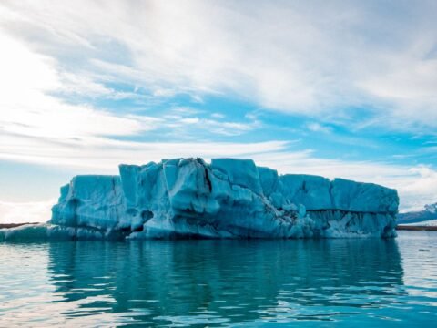 Large blue iceberg floating on calm water beneath a cloudy sky, illustrating concerns about rising global temperatures and ice loss