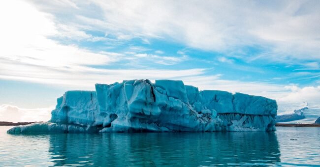 Large blue iceberg floating on calm water beneath a cloudy sky, illustrating concerns about rising global temperatures and ice loss