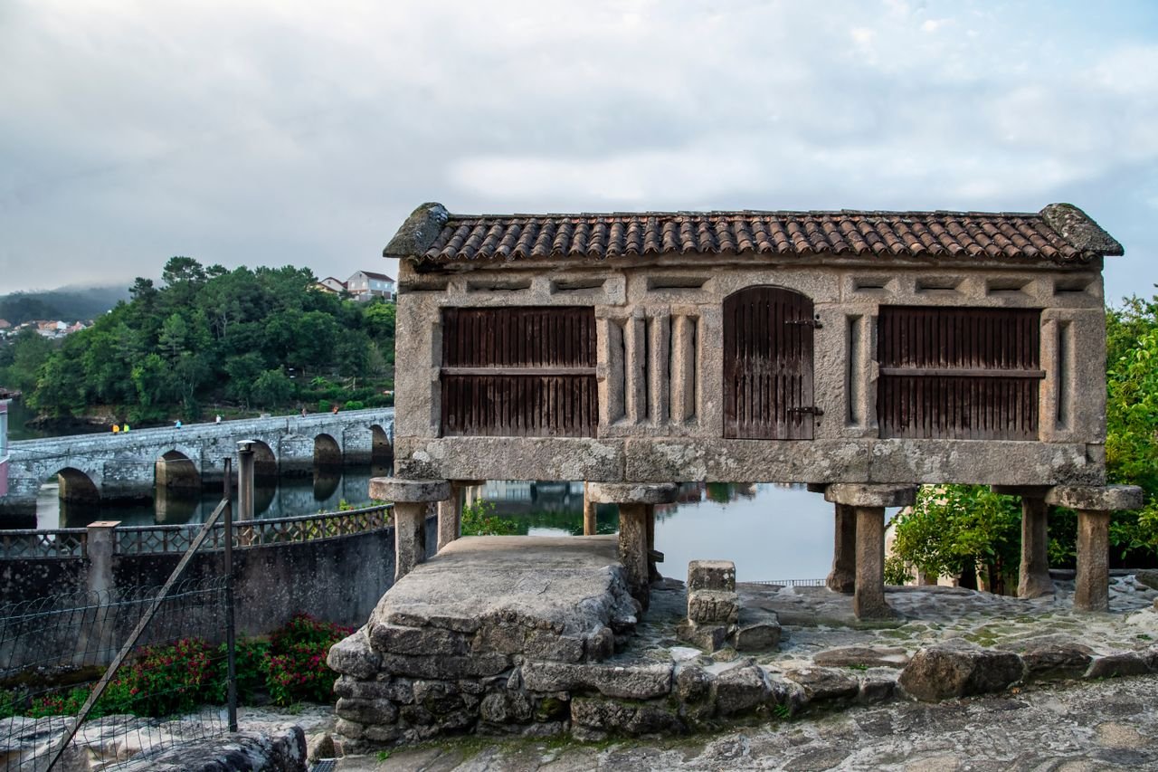 Traditional Galician hórreo raised on stone pillars beside a river and an old stone bridge