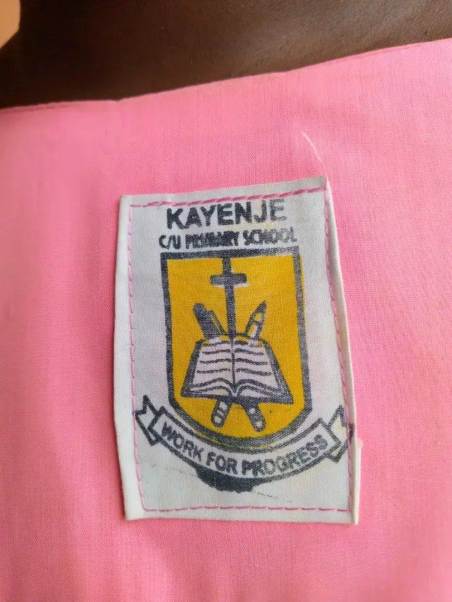 Embroidered badge of Kayenje Church of Uganda Primary School featuring a cross, an open book and the words Work for Progress.
