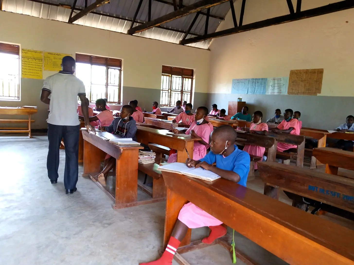 Teacher and pupils in a classroom with wooden desks at Kayenje Church of Uganda Primary School in Butambala, Uganda.