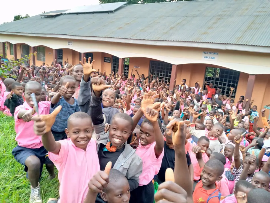Large group of smiling pupils in pink school uniforms raising their hands outside Kayenje Church of Uganda Primary School in Butambala, Uganda.